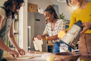 alt="Three people cooking together in a cozy kitchen—rolling dough, stretching it, and grating cheese—capturing the joy of collaborative meal prep." 