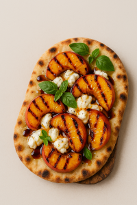 Closeup of grilled peach and burrata flatbread with hot honey drizzle, basil, and balsamic glaze on a rustic board
